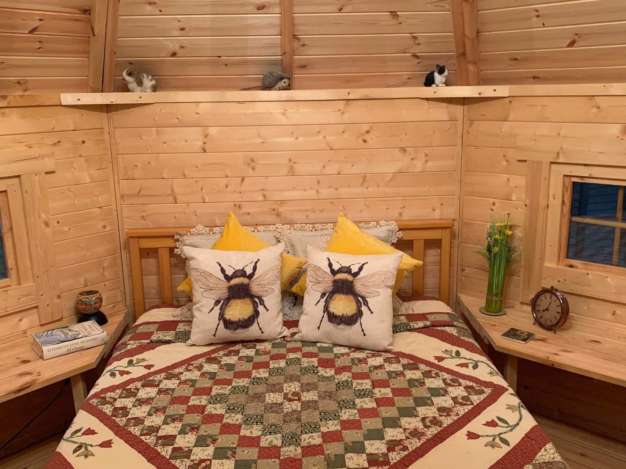 A cozy log cabin bedroom with a quilted bedspread, two pillows with bee designs, a wooden headboard, a bedside table with a book and a vase of flowers, and a clock.