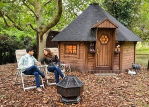 Two people sitting on chairs outside a wooden cabin with a hexagonal window and a small dog nearby. - Starry Meadow (Great Yarmouth)
