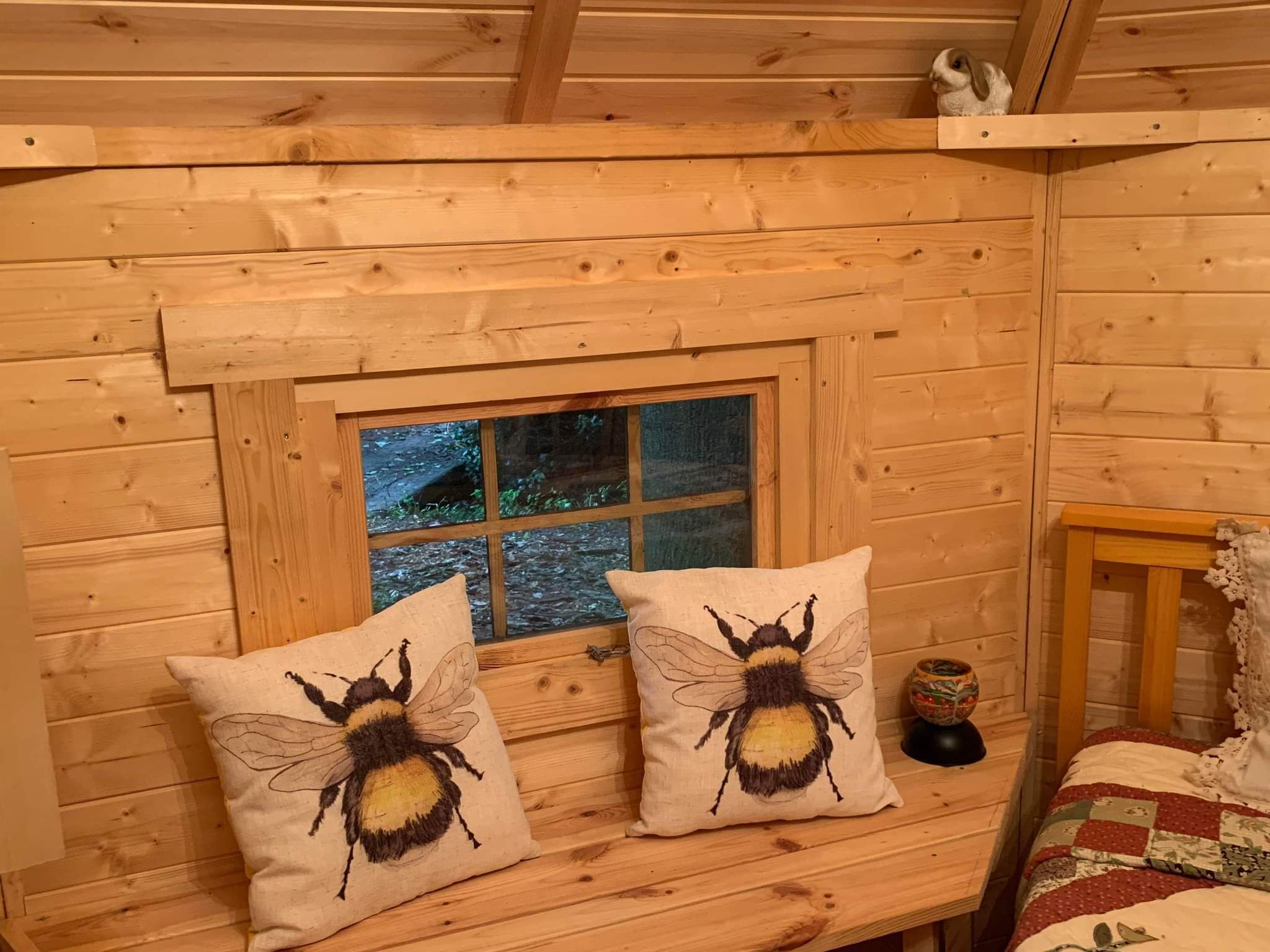 A cozy cabin room with wooden walls, a window with a view of a forest, two pillows with bee designs, and a quilted bedspread.