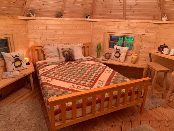 A cozy wooden cabin bedroom with a bed, pillows, a quilt, a bedside table with a lamp, a window, and a small table with a teapot and cups. - Starry Meadow (Great Yarmouth)