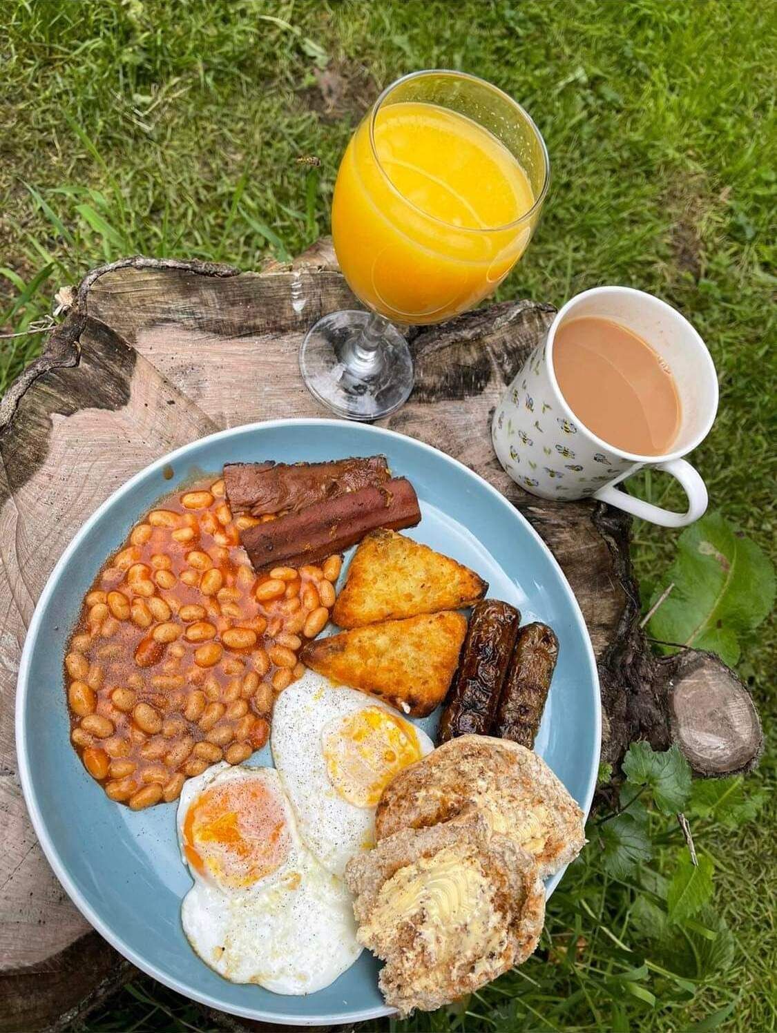 A plate of breakfast food including beans, sausages, fried eggs, and toast, with a glass of orange juice and a mug of tea on a wooden surface.