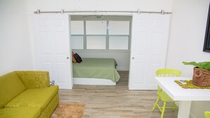 Habitació amb un llit, un sofà groc, una taula blanca amb caires i una finestra amb persianes.