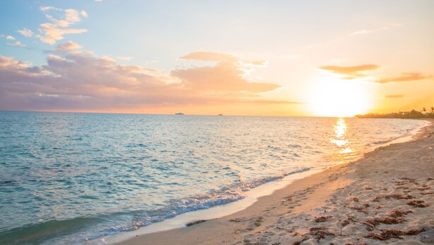 Une plage où l’on peut admirer le coucher de soleil sur l’océan, avec un rivage sablonneux et une terre lointaine à l’horizon.