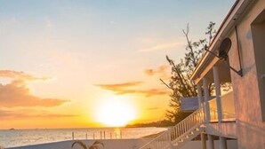 Une propriété située en bord de mer, dotée d’un escalier menant à un balcon avec vue sur la mer, une plage de sable et une vue dégagée sur le coucher du soleil.