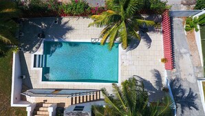 Vista aérea de una piscina rectangular con solárium azulejado, adyacente a un edificio con techo inclinado y una pequeña escalera que conduce a un nivel superior.