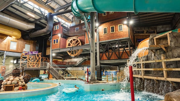 Parque acuático interior con una piscina grande, toboganes de agua y una zona de juegos de madera.