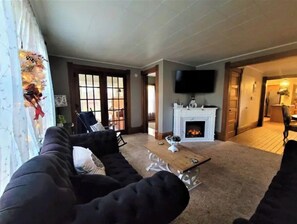A living room with a fireplace, a television mounted on the wall, and a large window with curtains. - Charming Farmhouse in the Orchards near Lake Mich! (New Era)