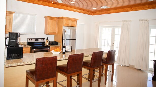 A kitchen with wooden cabinets, a stainless steel refrigerator, a stove, and a coffee maker. There are four wooden chairs around a central island. - Tangerine Sunsets Satsuma One (Nassau)