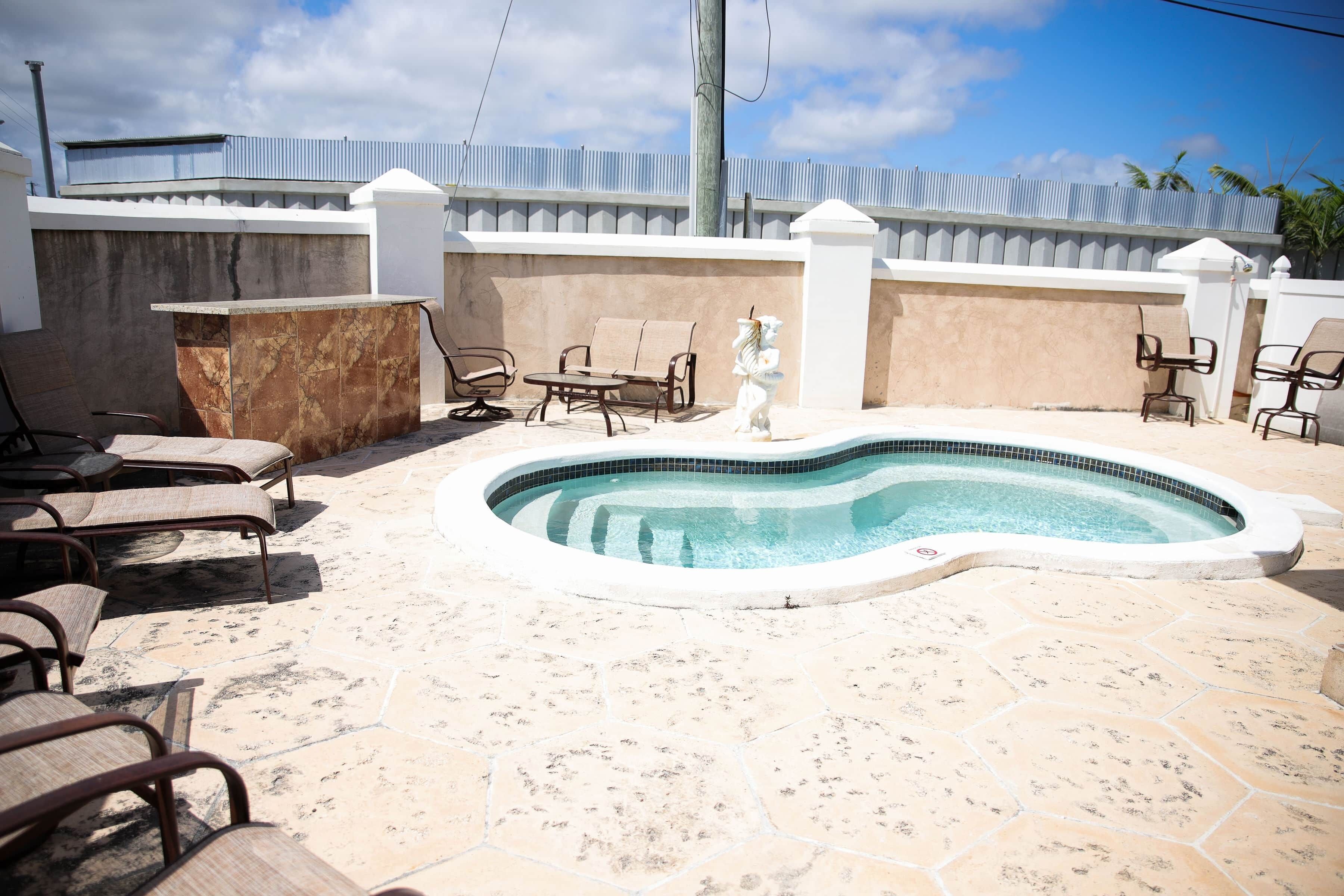 Piscina com espreguiçadeiras, deck de pedra e parede branca com cerca de metal.