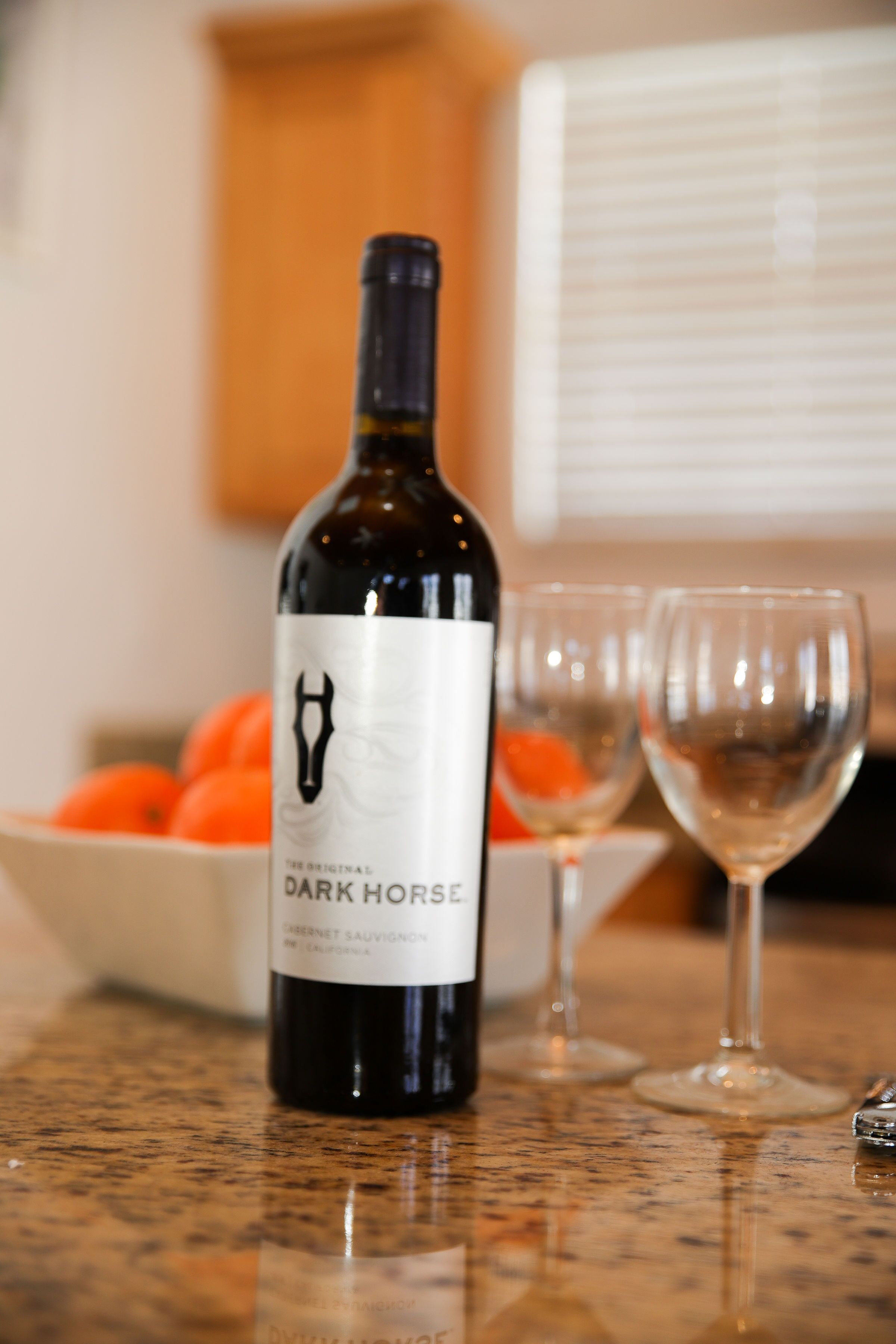 Una botella de vino Dark Horse sobre una encimera de cocina, junto a un bol de naranjas y dos copas de vino.