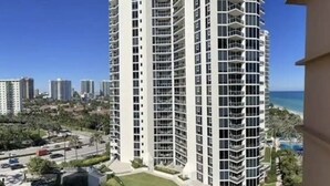 A high-rise building with multiple balconies, surrounded by greenery and a parking area.