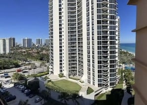 A high-rise building with multiple balconies, surrounded by greenery and a parking area.