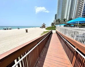 A wooden boardwalk leading to a beach with a clear view of the ocean and tall buildings in the background.