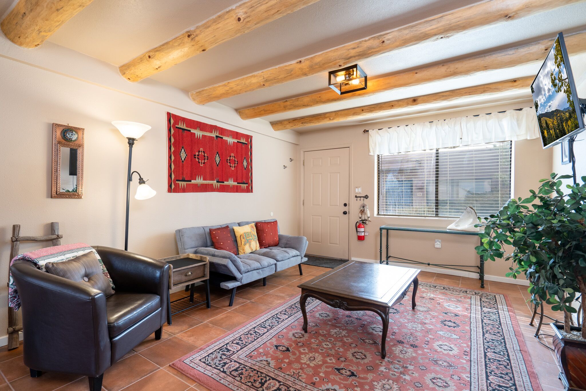 A living room with a leather sofa, a wooden coffee table, a red patterned rug, a wall-mounted TV, and a large window with blinds.