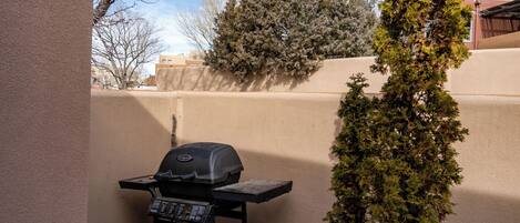 A backyard with a barbecue grill, a small wooden table, and a tall evergreen tree.