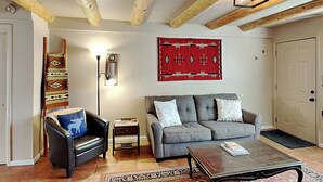 A living room with a grey sofa, a black armchair, a coffee table, a patterned rug, a wall-mounted lamp, and a red decorative wall hanging. - Casita De Alces by Kokopelli Property Management (Santa Fe)
