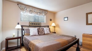 A neatly arranged bedroom with a bed, bedside lamps, a nightstand, a dresser, and a window with blinds. - Casita De Alces by Kokopelli Property Management (Santa Fe)