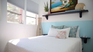 A bedroom with a bed, a nightstand, a window, and a shelf with a painting and a plant. - Tangerine Sunsets Castaway, Tangerine Sunsets Castaway (Nassau)