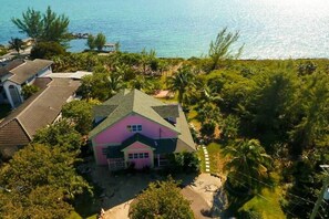 An aerial view of a pink house with a green roof surrounded by lush greenery and a clear blue sea.