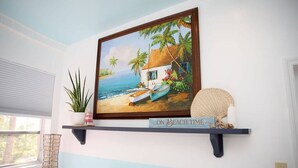 A room with a painting of a beach scene, a shelf with decorative items, and a plant. - Tangerine Sunsets Castaway, Tangerine Sunsets Castaway (Nassau)