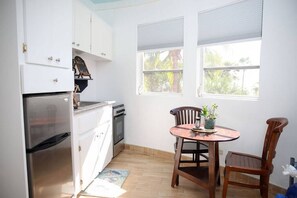 A compact kitchen with white cabinets, a stainless steel refrigerator, a stove, and a small dining table with two chairs.