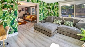 Una sala de estar con un sofá modular, una mesa de centro y una pared con estampado de hojas tropicales. Se ve una ventana con vistas al exterior.