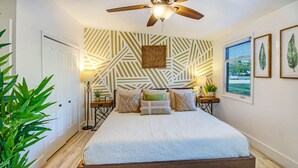 Un dormitorio con cama, ventilador de techo, pared con estampado y ventana con persianas.