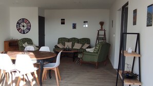 A dining area with a round wooden table and white chairs, a sofa with patterned cushions, and a wall clock.