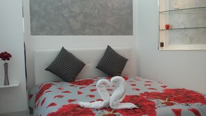 Een slaapkamer met een bed, overdekt met een bloemenlaken, twee kussens en een nachtkastje met een rode bloem in een vaas.