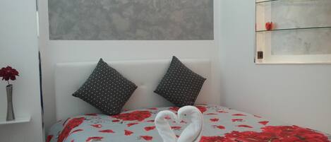 Une chambre à coucher avec un lit recouvert d’une couvre-lit à motifs floraux, deux oreillers et un vase contenant une fleur rouge, posé sur une table de chevet.