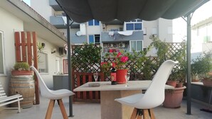 Een terras buiten met een tafel en twee witte stoelen, een plant in pot en een fles wijn.