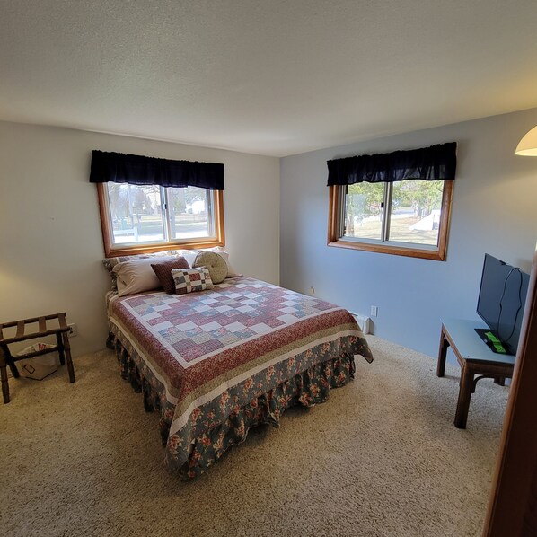 A bedroom with a large bed, two windows with black valances, a desk with a chair, and a television. - Marina View in the Village (Pentwater)