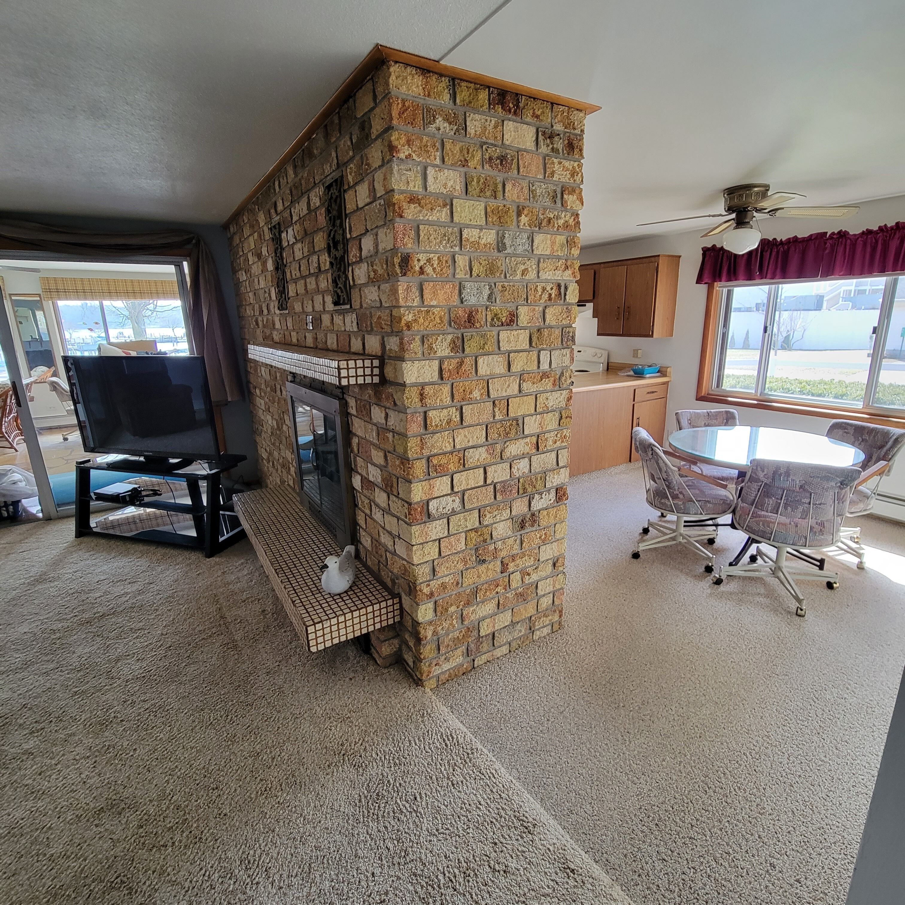 A living room with a brick fireplace, a dining area with a table and chairs, and a kitchen area with wooden cabinets and a window.