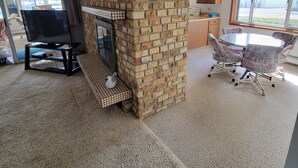 A living room with a brick fireplace, a dining area with a table and chairs, and a kitchen area with wooden cabinets and a window.