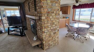 A living room with a brick fireplace, a dining area with a table and chairs, and a kitchen area with wooden cabinets and a window.
