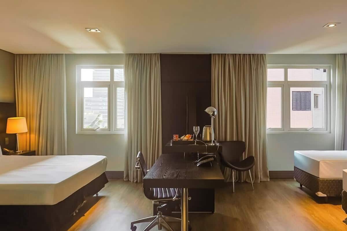 A hotel room with two beds, a desk, a chair, and a lamp.