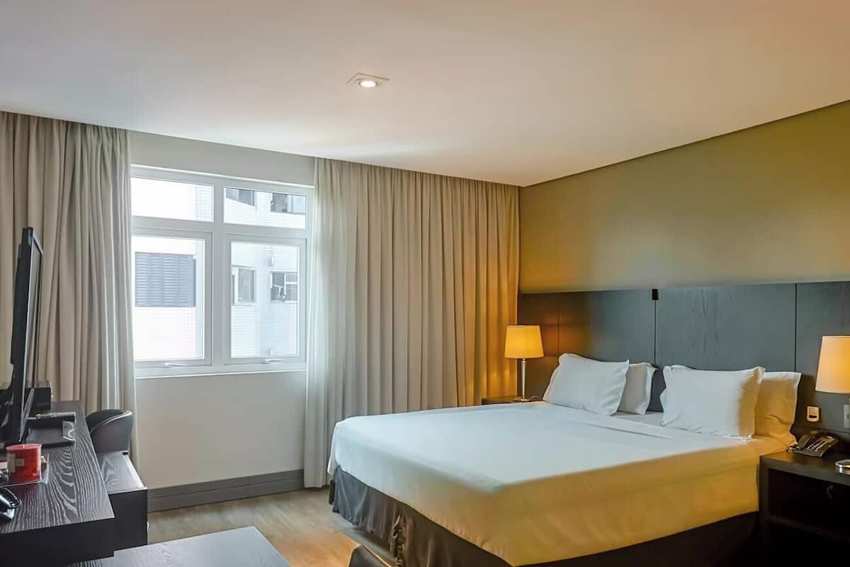 A hotel room with a bed, bedside tables with lamps, a desk with a computer, a chair, and a window with curtains.