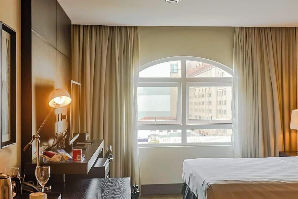A hotel room with a bed, two bedside lamps, a desk with a lamp, a chair, and a view of buildings through a window.