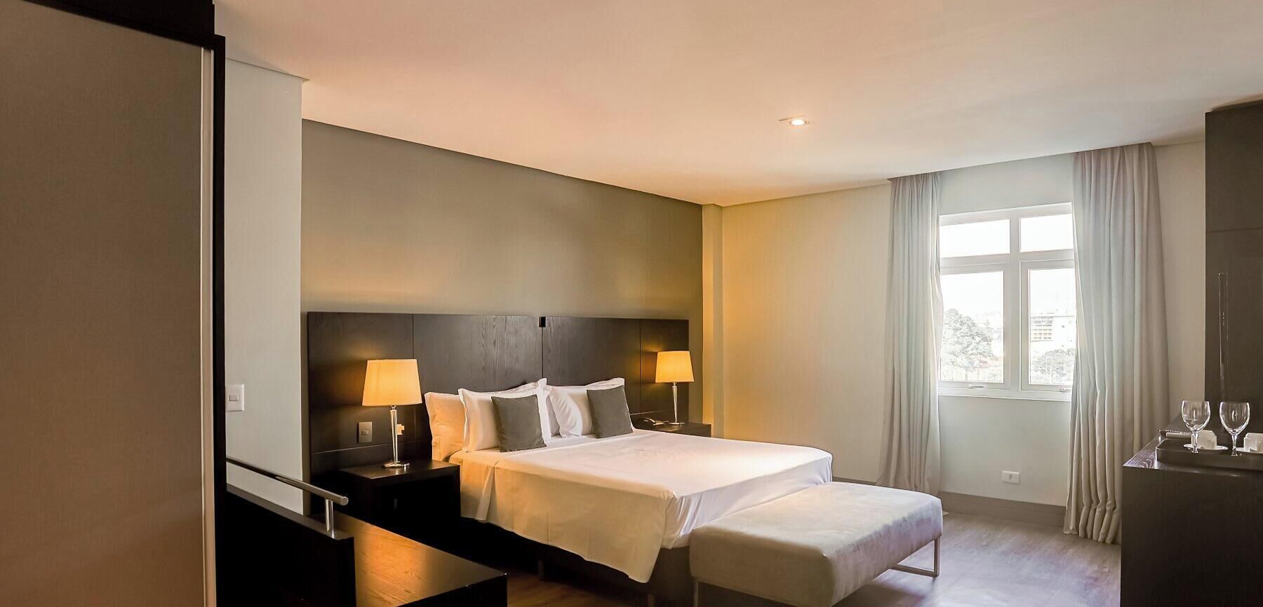 A hotel room with a bed, bedside tables, a desk with a chair, and a window with curtains.