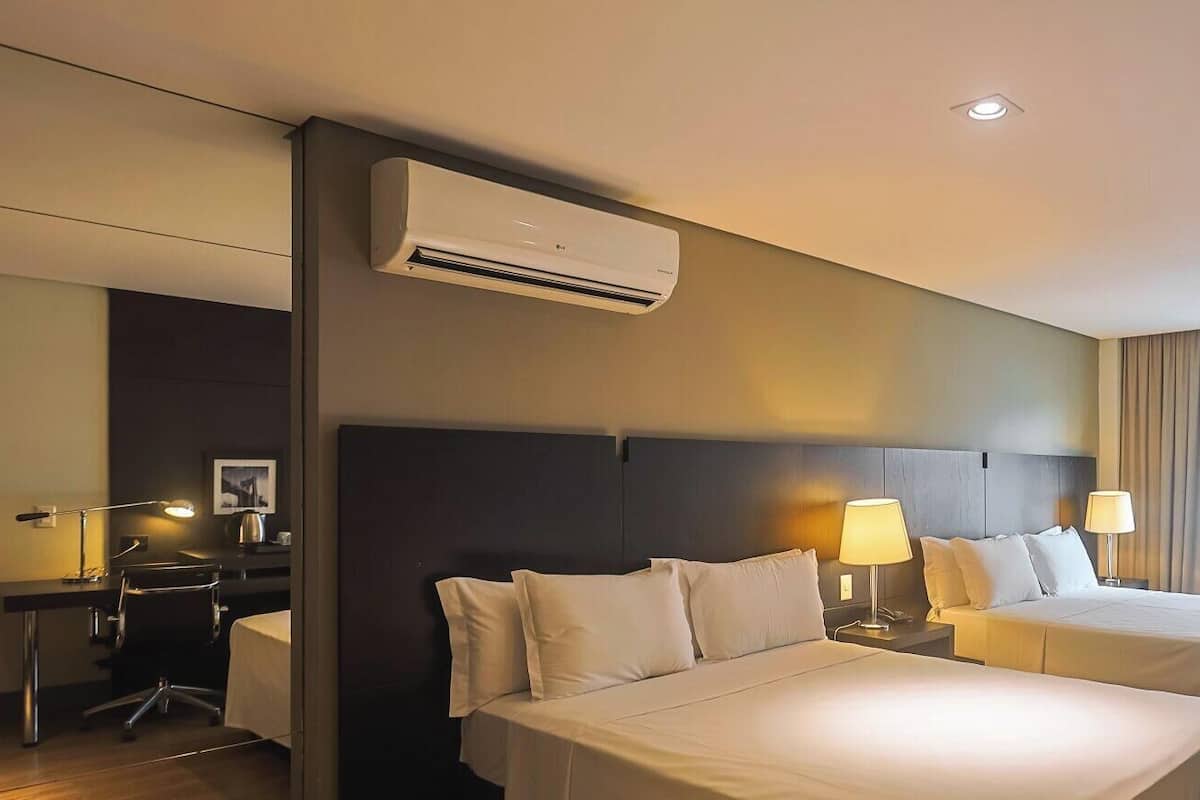 A hotel room with two beds, a desk, a chair, an air conditioner, and a window with curtains.