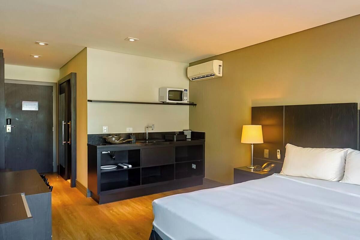 A hotel room with a bed, bedside tables, a desk, a microwave, a phone, and a small kitchenette.