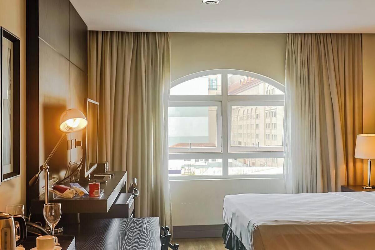 A hotel room with a bed, bedside lamps, a desk with a lamp, a coffee maker, and a view of buildings through a window.