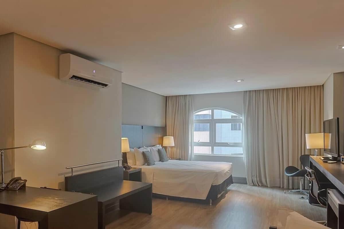 A hotel room with a bed, desk, and chair, a window with curtains, and an air conditioning unit.
