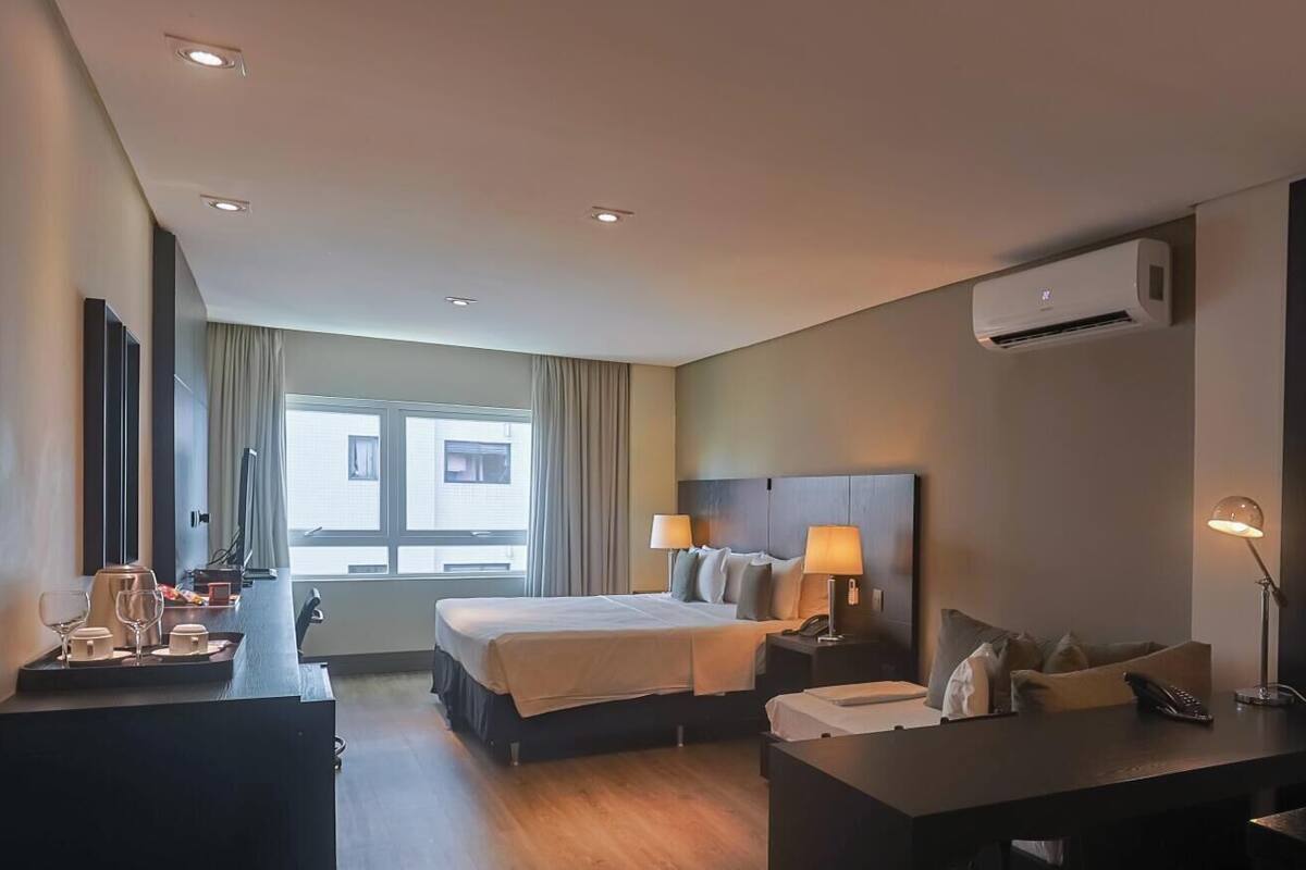 A hotel room with a bed, bedside tables, a desk, a chair, an air conditioning unit, and a window.