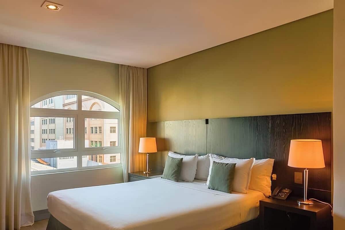 A hotel room with a bed, two bedside lamps, a nightstand, and a view through a window.