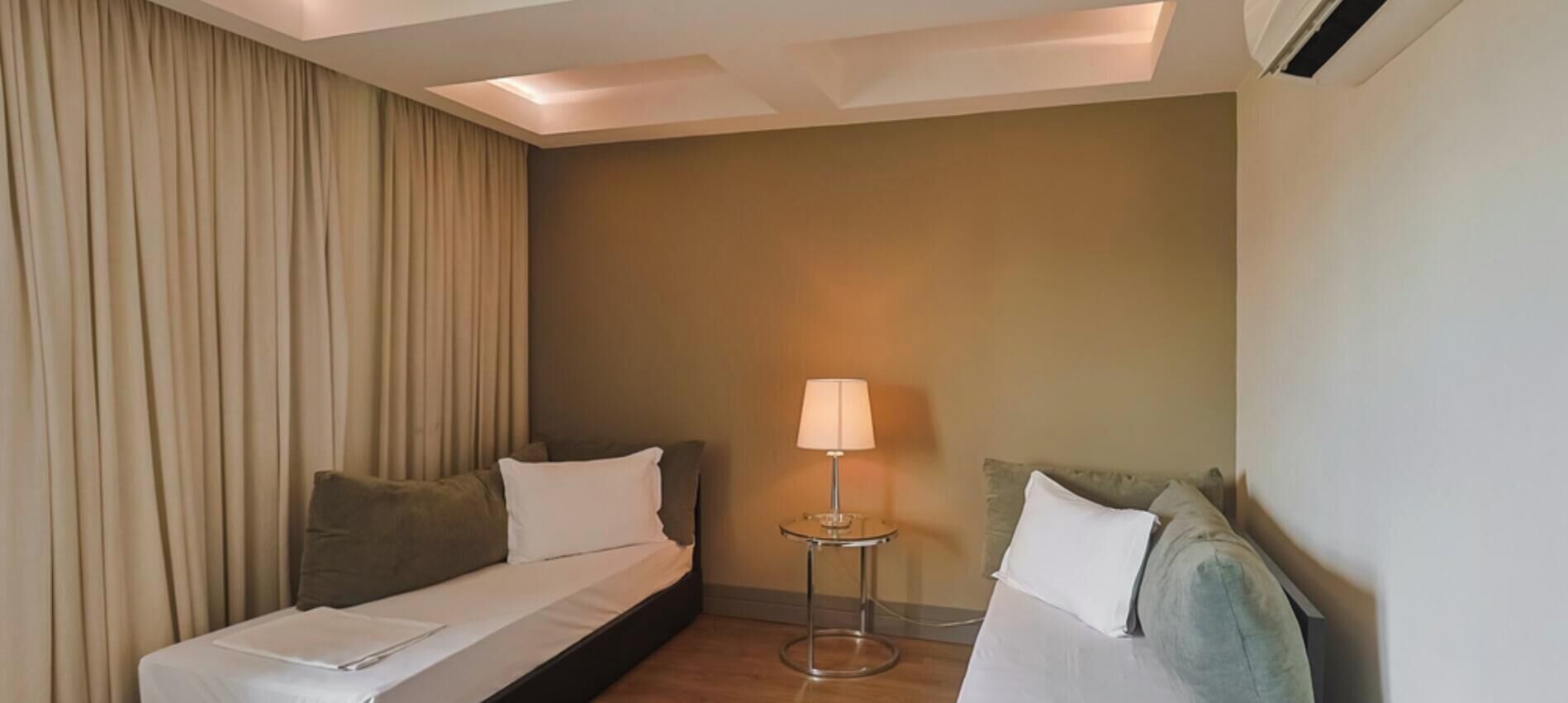 Una habitación de hotel con dos camas, una mesita de noche con lámpara y un amplio ventanal con cortinas.
