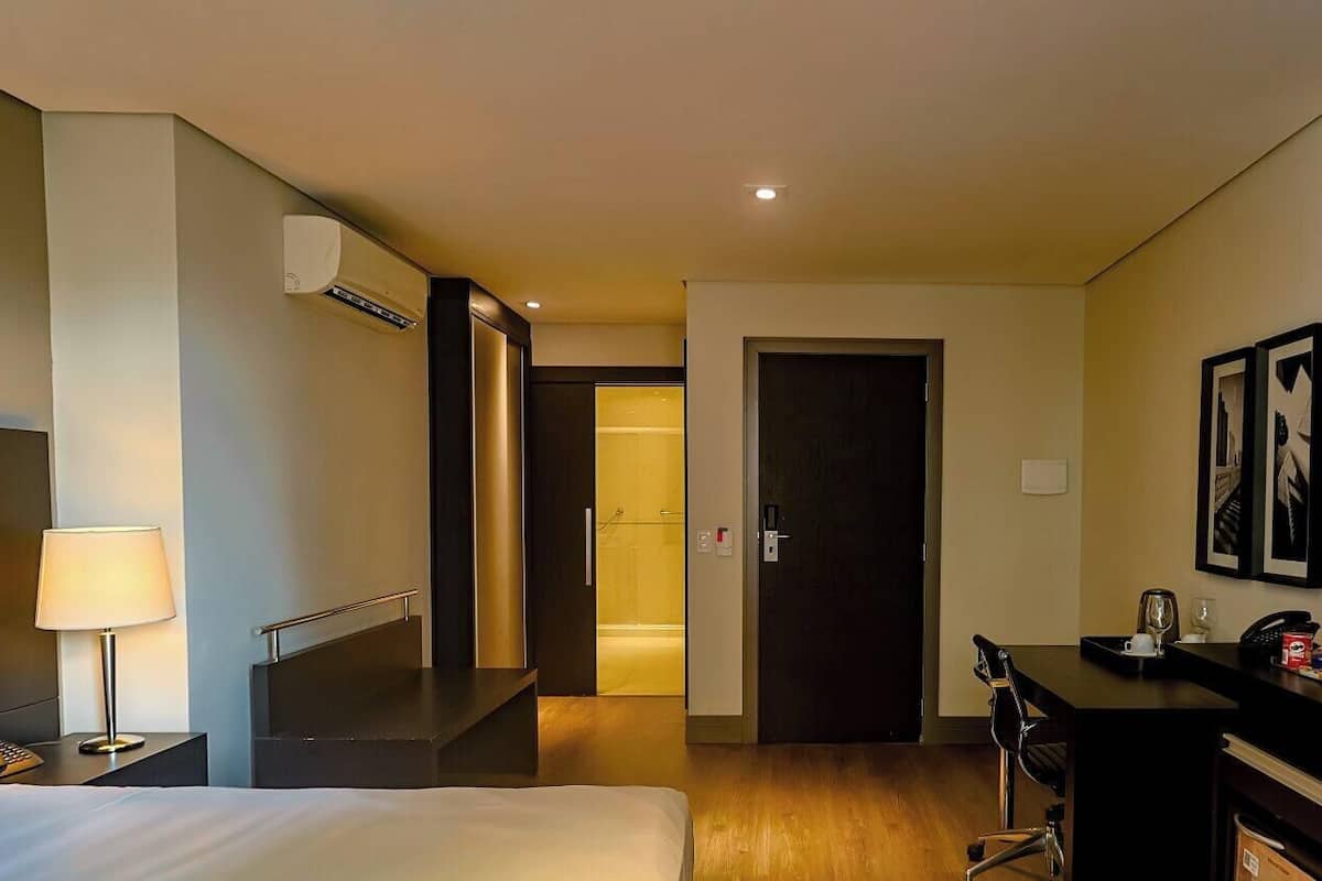 A hotel room with a bed, bedside table, lamp, TV, desk, chair, and a bathroom visible through an open door.