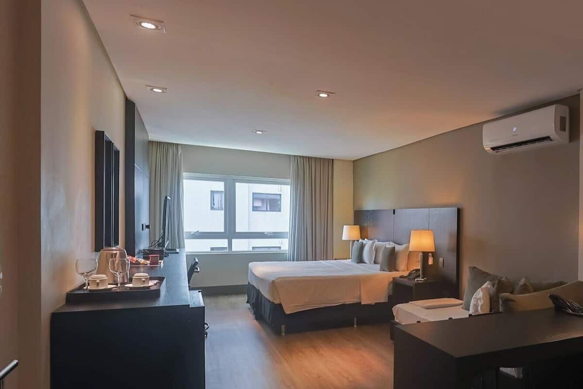 A hotel room with a bed, bedside tables, a desk, an air conditioning unit, and a window with curtains.
