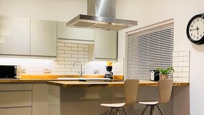 A modern kitchen with a stainless steel range hood, a sink, and a coffee maker. There are two bar stools and a clock on the wall. - Luxury 2 bed Apartment Golden Triangle w Parking (Norwich)