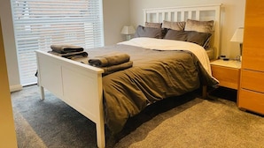 A bedroom with a bed, a nightstand, a dresser, and a window with blinds. - Luxury 2 bed Apartment Golden Triangle w Parking (Norwich)